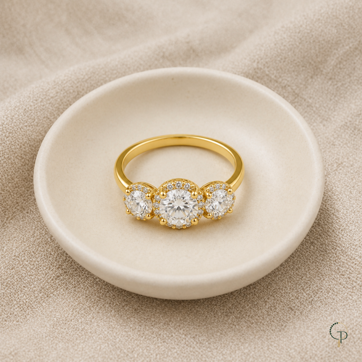 Gold-color three-stone halo ring arranged on a neutral ceramic dish