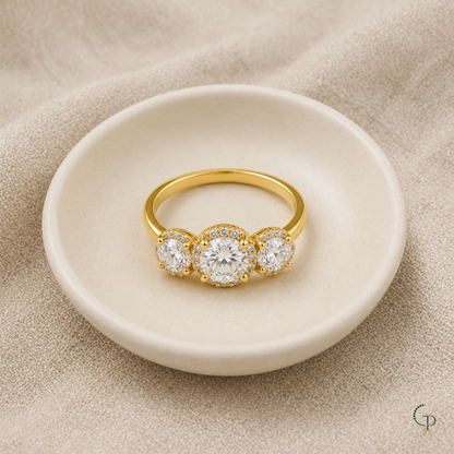 Gold-color three-stone halo ring arranged on a neutral ceramic dish