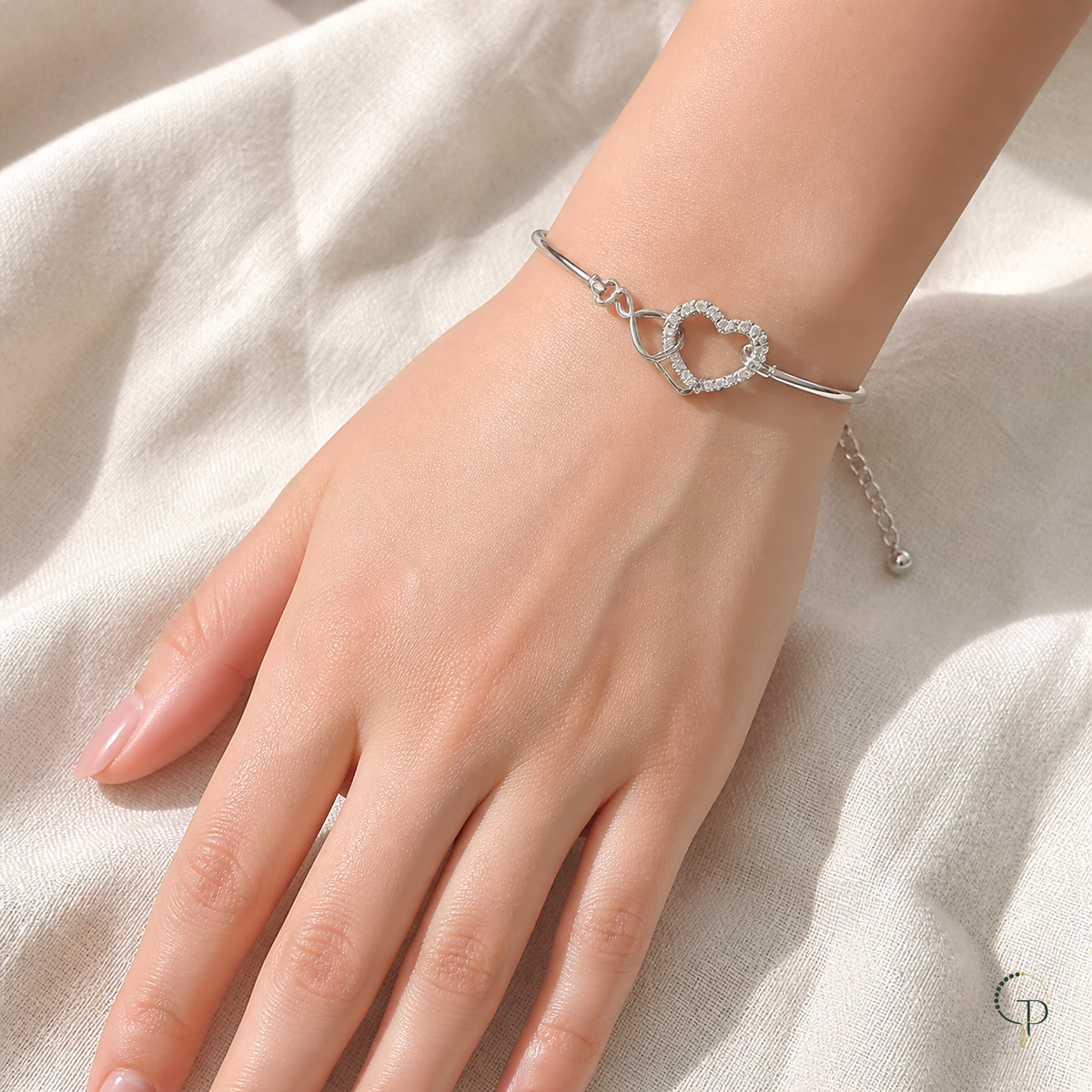 Interlocking double-heart bracelet shown at wrist scale over ivory fabric