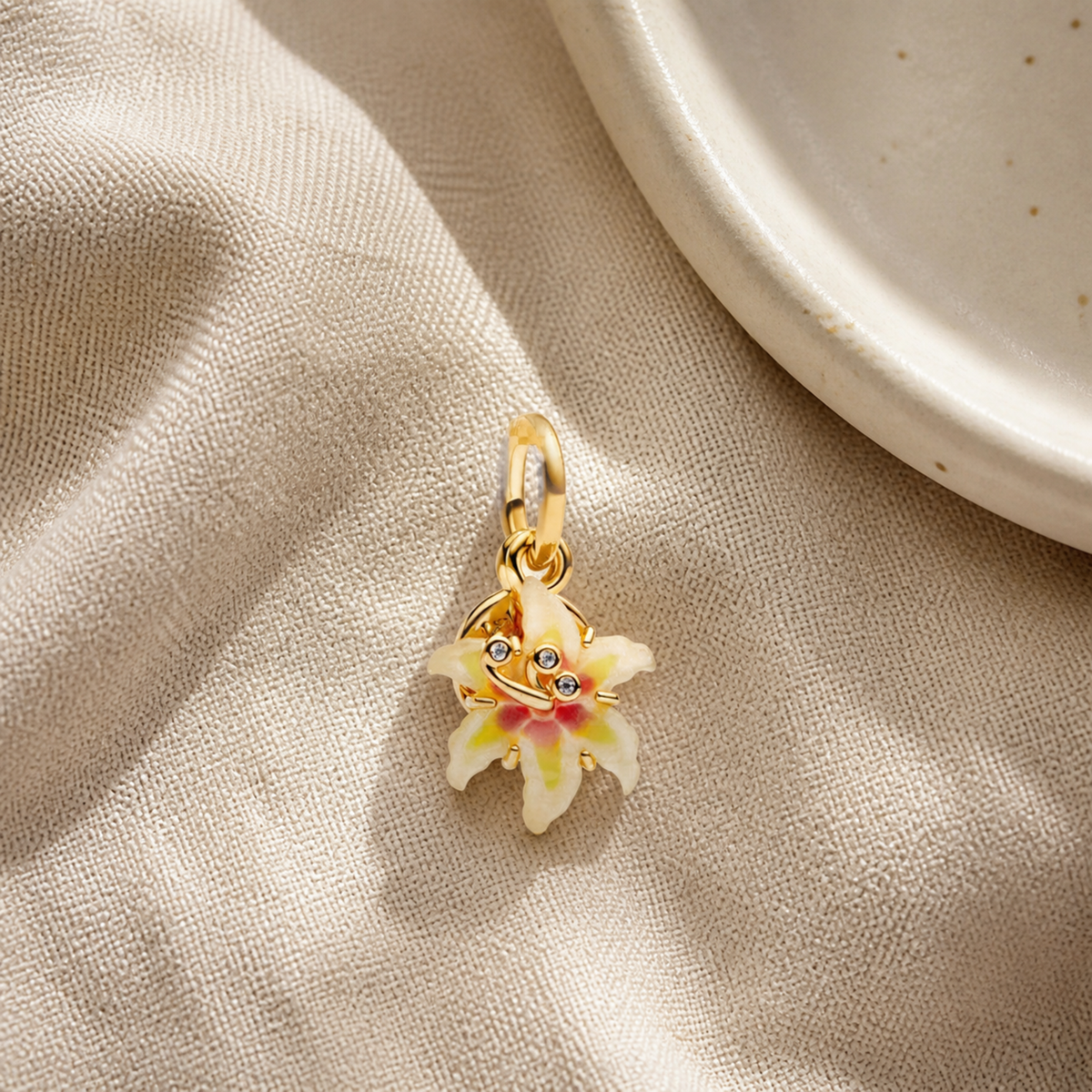 Enamel flower pendant charm styled on oatmeal linen beside a ceramic dish