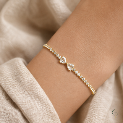 Gold-color twin heart bow tennis bracelet worn on a wrist with an oatmeal linen sleeve
