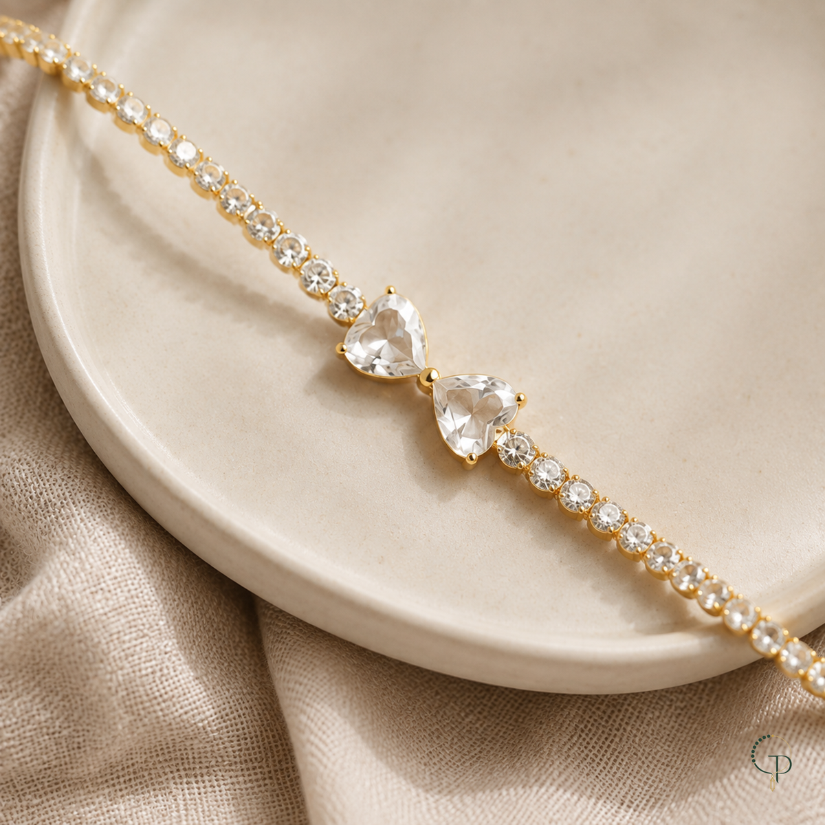 Gold-color twin heart bow tennis bracelet styled across an ivory ceramic dish