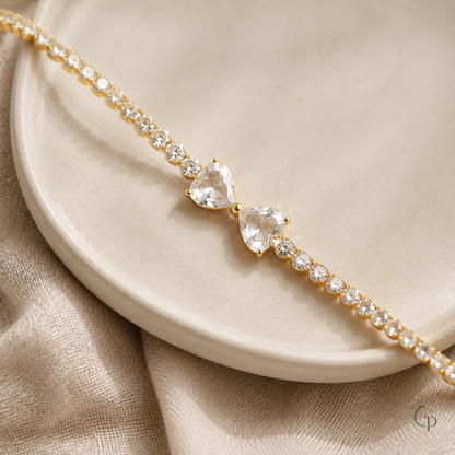 Gold-color twin heart bow tennis bracelet styled across an ivory ceramic dish